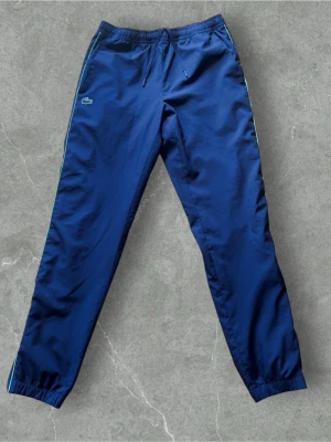 Lacoste Track Pants - Stilrena Lacoste track pants i mörkblå färg med diskret krokodillogga och kontrastdetaljer längs sidorna. Tillverkade i ett lätt och bekvämt material med elastisk midja och dragsko för optimal passform.  Skick: Nyskick, utan anmärkningar. Passform: Avsmalnande modell med elastiska benslut. Perfekta för både vardag, träning och en sportig streetwear-look.
