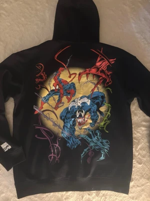 Svart Spider-Man hoodie med tryck - Cool svart hoodie med stor färgglad Marvel tryck på ryggen med Spiderman och venom. Framsidan har 'The Amazing Spiderman logga och magficka. Mjuk bomullskänsla och huva för extra stilpoäng. Har aldrig haft på mig den.