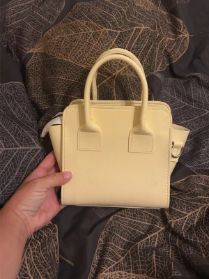 Ljusgul minihandväska med handtag - Söt ljusgul minihandväska i slätt konstläder med två stadiga handtag och avtagbar axelrem. Väskan har en stilren och boxig form som ger en trendig look. Perfekt för dig som vill ha en liten och smidig väska till mobilen och det viktigaste.
