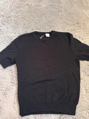 Svart stickad kortärmad topp - En enkel och stilren svart stickad topp med korta ärmar och rund halsringning. Perfekt att matcha med jeans eller kjol för en clean look. Mjuk och bekväm med ribbade kanter vid ärmslut och nederkant.