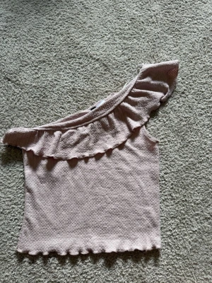 Ljusrosa oneshoulder topp med volang - Superfin ljusrosa topp i oneshoulder-modell med volang längs halsringningen. Toppen har en vågig kant nertill och är gjord i ett mjukt, strukturerat material. Perfekt för dig som vill ha en söt och trendig look. Liten i storleken – skulle rekommendera den för någon som vanligtvis har en storlek mindre.