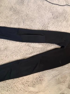 Svarta leggings med ficka - Säljer ett par svarta leggings med en praktisk ficka på benet. De har en tight passform och är perfekta för en avslappnad stil. Materialet ser stretchigt och bekvämt ut, vilket gör dem smidiga att röra sig i.