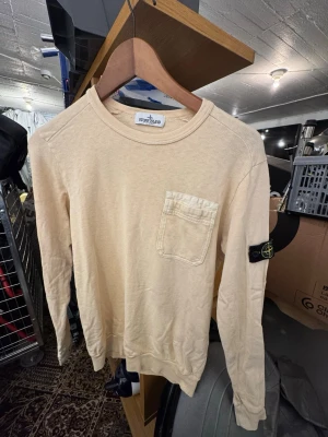 Beige långärmad tröja Stone Island - Beige långärmad tröja från Stone Island med rund halsringning och bröstficka. Tröjan har en klassisk svart logotyp-patch på vänster ärm och ribbade muddar vid ärmslut och nederkant. Perfekt för en avslappnad och stilren look. Finns att hämta i Gröndal. Junior så Small är passformen. Nypris är 2699kr.