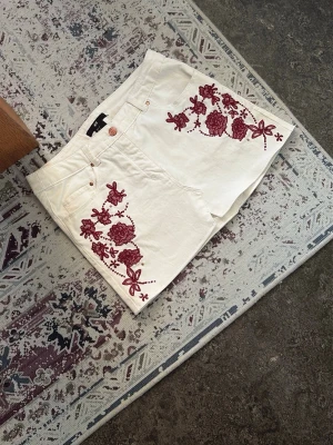 Vita shorts med röd brodyr - Snygga vita shorts med röda blommiga broderier på båda sidor framtill. Klassisk femficksmodell med knapp och dragkedja. Shortsen har en rak passform och är perfekta för sommarens alla äventyr.