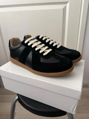Maison Margiela Svarta 43 - Stilrena svarta sneakers med beige sula och vita snören. Ovandelen är i en mix av mocka och slätt läder för en schysst kontrast. Insidan är ljus och sulan är i klassisk gummifärg. Perfekta för dig som gillar minimalistisk och tidlös stil.