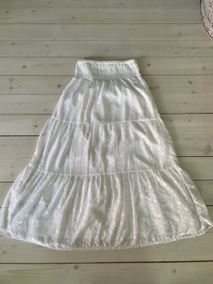 Vit långkjol från Gina Tricot - Luftig vit långkjol från Gina Tricot med bred resår i midjan och diskret mönstrad struktur i tyget. Kjolen har flera volanger som ger en flowy känsla och passar perfekt till varma dagar.