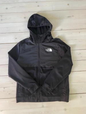 Svart hoodie från The North Face - Snygg svart hoodie från The North Face med dragkedja framtill och vit logga på bröstet. Hoodien har huva och två fickor på sidorna. Tillverkad i ett mjukt och lite glansigt material som känns sportigt och modernt. Perfekt för dig som gillar streetwear. Funkar lika bra för kvinnor som män. Lite nopprig men går att fixa