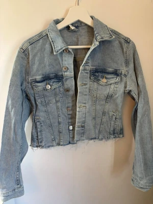 Ljusblå croppad jeansjacka - Säljer en väldigt fin ljusblå jeansjacka med croppad passform och rå kant nertill. Klassisk krage, två bröstfickor med lock och knappar samt långa ärmar. Perfekt för en avslappnad och cool stil.