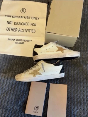Golden Goose vita sneakers med glitter - Golden Goose sneakers i vitt läder med en glittrig guldstjärna på sidan och svart glitter på hälen. Skorna har grå skosnören och en klassisk låg siluett. Snygg och unik design som sticker ut i mängden. Köpte av en på Plick, men dom passar inte så säljer vidare.💗