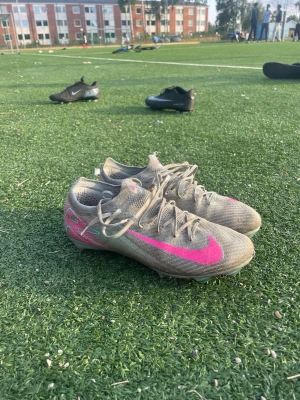Beige Nike fotbollsskor med rosa swoosh - Säljer ett par beige Nike fotbollsskor med en stor rosa swoosh på sidan. Skorna har snörning och låg profil, designade för spel på konstgräs. Tillverkade i syntetmaterial för bra grepp och känsla på planen. Perfekta för dig som vill sticka ut på matchen!