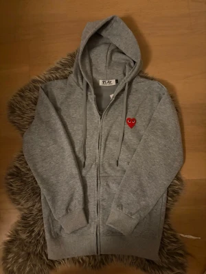 Grå PLAY zip hoodie S - Säljer en grå zip-hoodie / kofya från Comme des Garçons PLAY med det ikoniska röda hjärtat med ögon broderat på bröstet. Hoodien har dragkedja, två fickor fram, justerbar huva med snören och ribbade muddar. Perfekt för en avslappnad och trendig stil. Endast testad. 