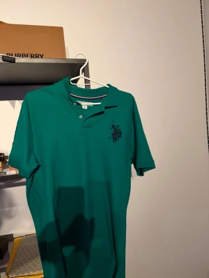 Grön pikétröja med tryck - Snygg grön pikétröja med korta ärmar och klassisk krage. Tröjan har knappar vid halsen och ett mörkare tryck på bröstet. Perfekt till jeans eller shorts för en avslappnad look.