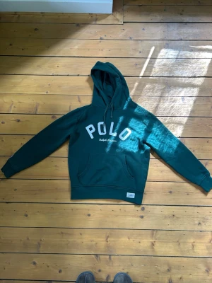 Grön hoodie från Polo Ralph Lauren - Mörkgrön hoodie med stor huva och magficka från Polo Ralph Lauren. Stor vit POLO-text framtill och dragsko i huvan. Klassisk och avslappnad passform, perfekt till jeans eller shorts.  Storlek S, tveka inte att ställa frågor. 