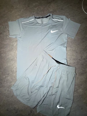 Ljusgrå Nike Miller set - Säljer ett ljusgrått träningsset från Nike med t-shirt och shorts. T-shirten har korta ärmar, rund hals och reflekterande detaljer på axlarna samt Nike-logga på bröstet. Shortsen har resår i midjan, dragsko och reflekterande Nike-logga. Perfekt för löpning eller gym.