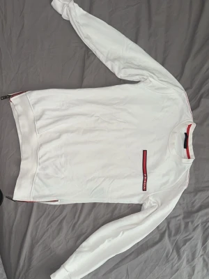 Vit Prada tröja  - Snygg vit sweatshirt med lång ärm och rund hals. Tröjan har en röd och svart randig detalj vid halsen och en röd rand med text på bröstet. Perfekt för en clean och sportig look. Materialet känns mjukt och bekvämt. Storlek S passar mig på 186cm och 78kg