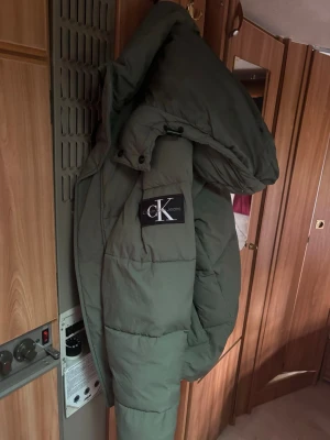 Grön pufferjacka från Calvin Klein - Säljer en grön pufferjacka från Calvin Klein Jeans med stor huva och tydlig logga på armen. Perfekt för kalla vinterdagar och riktigt snygg. 