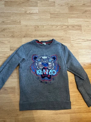 Grå Kenzo sweatshirt med tigertryck - Säljer en grå sweatshirt från Kenzo med det ikoniska broderade tigerhuvudet och texten 'KENZO PARIS' framtill. Tröjan har rund halsringning, långa ärmar och är gjord i mjukt material. Perfekt för dig som vill sticka ut med en cool streetstyle-look.
