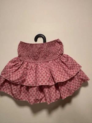 Rosa volangkjol från MEET x TIGER - Supersöt kort kjol i rosa med vitt blommönster från MEET x TIGER. Kjolen har bred smock i midjan och dubbla volanger och ett stretchigt material. Kjolen har inbygda shorts.