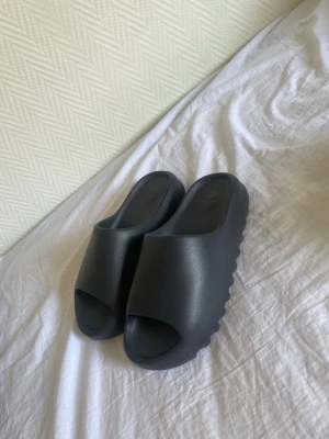 Svarta slides från Adidas - Säljer ett par svarta slides från Adidas med chunky sula och öppen tå. Tillverkade i ett mjukt och flexibelt syntetmaterial som ger skön komfort. Perfekta för chill dagar hemma eller till stranden.