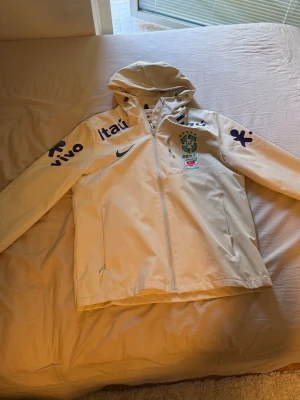 Vit Brasilien Nike vindjacka med huva - Storlek L men den passar egentligen S/M!! Snygg vit vindjacka från Nike med Brasilien-logga och sponsormärken i blått. Jackan har huva, hel dragkedja och meshfoder på insidan. Perfekt för dig som gillar sportig stil och vill sticka ut med landslagskänsla.