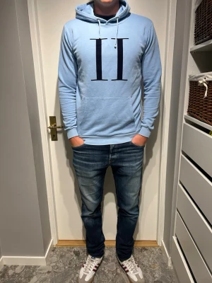 Ljusblå hoodie med tryck - Snygg ljusblå hoodie med stor svart romersk siffra II tryckt på bröstet. Hoodien har huva med vita dragsnören och en klassisk magficka framtill. Tillverkad i mjukt material och har ribbade muddar vid ärmar och nederkant. Perfekt för en avslappnad stil. Litet hål vid fickan men inget man ser.