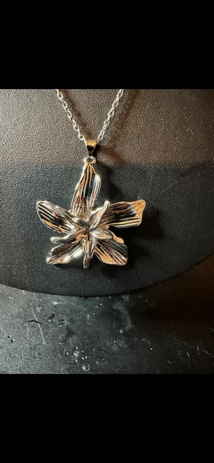 Silverfärgat halsband med stor blomma - Unikt halsband med en stor, silverfärgad blomma som hänge. Blomman har detaljerade blad och en glänsande finish. Kedjan är tunn och matchar hängets metalliska look. Perfekt för dig som vill sticka ut med ett statement-smycke.