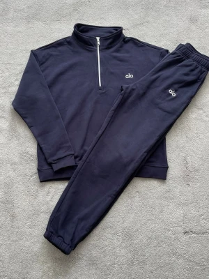 Mörkblå loungewear-set från alo - Snyggt och bekvämt loungewear-set från alo i mörkblått. Setet består av en långärmad tröja med halv dragkedja och matchande byxor med resår i midjan och benslut. Båda delarna har alo-logga framtill. Perfekt för chill dagar hemma.