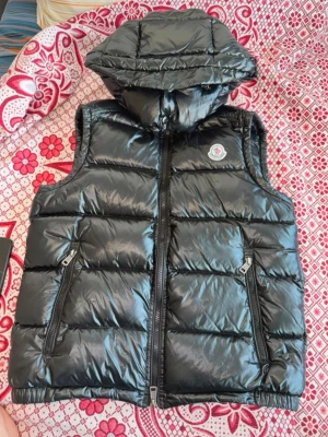 Svart dunväst med huva Moncler - Snygg svart dunväst från Moncler med glansig finish och avtagbar huva. Västen har två dragkedjeförsedda fickor framtill och klassisk Moncler-logga på bröstet. Perfekt för lager-på-lager och streetstyle.