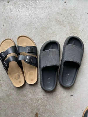 Två par svarta sandaler - Säljer två par sandaler: ett par med två justerbara remmar och spännen i svart läderimitation dessa är helt nya. samt ett par slip-in sandaler i helsvart syntetmaterial med bred rem från Lager 157 Båda paren har öppen tå och är perfekta för sommaren.