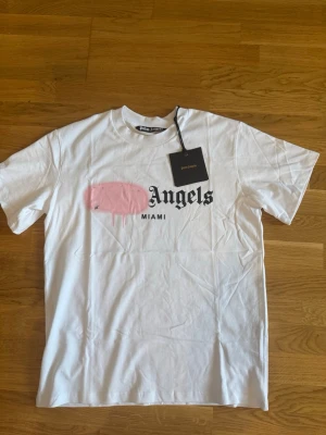 Vit Palm Angels t-shirt med tryck - Säljer en vit t-shirt från Palm Angels med svart text och rosa grafiskt tryck på bröstet. Modellen har klassisk passform, rund hals och korta ärmar. Materialet är mjuk bomull och t-shirten har en streetwear vibe.
