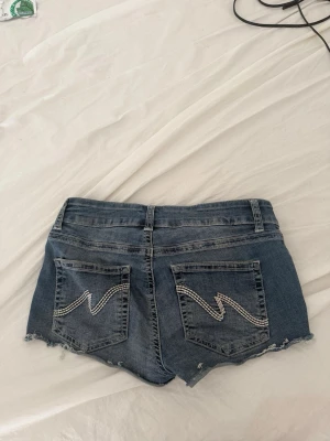 Nelly lågmidjade jeansshorts cutoff fransade Y2K blå - Snygga jeansshorts från Nelly med dekorativa sömsticksdetaljer på bakfickorna — klassisk Y2K-känsla! Fransade kanter, lågmidjad passform — ser ut som en het sommaroutfit med ett linne eller crop top. Perfekta för festival, semester eller en varm sommardag. Begagnat skick, inga synliga defekter förutom de fransar som hör till stilen.