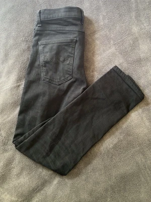 Svarta nudie jeans - Säljer ett par svarta nudie jeans med klassisk femficksdesign och smal passform. Jeansen har silverfärgade nitar och knapp, samt normalhög midja. Perfekta för dig som gillar en stilren och enkel look.