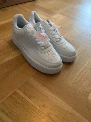 Nike Air Force 1 vita sneakers - Klassiska vita Nike Air Force 1 sneakers i lågt utförande. Skorna har ovandel i slätt läder, perforerad tåbox för ventilation och vit sula. Diskret Nike-logga på sidan och AIR-detalj bak. Perfekta för en clean och stilren look.