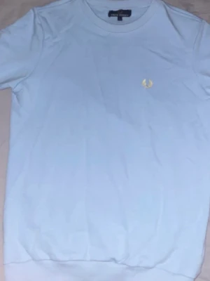 Fred Perry sweatshirt - Säljer en ljusblå sweatshirt från Fred Perry med klassisk gul logga broderad på bröstet. Sweatshirten har rund hals och korta ärmar, tillverkad i mjuk bomull för skön känsla. Perfekt till jeans eller shorts för en clean look.