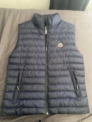 Moncler väst - Snygg mörkblå dunväst från Moncler med quiltad design och hög krage. Västen har två dragkedjeförsedda fickor och Moncler-logga på bröstet. Perfekt för lager-på-lager och håller dig varm under kyliga dagar. Inga skador eller hål. Storlek S