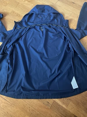 Mörkblå regnjacka från Nike - Snygg mörkblå regnjacka från Nike med huva och hel dragkedja framtill. Jackan har två sidofickor med dragkedja och ett diskret Nike-märke på bröstet. Tillverkad i ett lätt och vattenavvisande material som passar perfekt för regniga dagar.