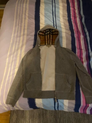 Grå hoodie från Burberry med dragkedja - Säljer en grå hoodie från Burberry med dragkedja och två fickor framtill. Huvan har det klassiska Burberry-mönstret på insidan och dragsko. Materialet är mjukt och skönt, perfekt för chill dagar. Broderad Burberry-logga på bröstet.