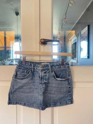 Kort jeanskjol i ljusblå denim - Säljer en klassisk kort jeanskjol i ljusblå denim med fem fickor och bälteshällor. Kjolen har en rak passform och stängs med knapp och dragkedja framtill. Perfekt basplagg för en avslappnad och trendig look.