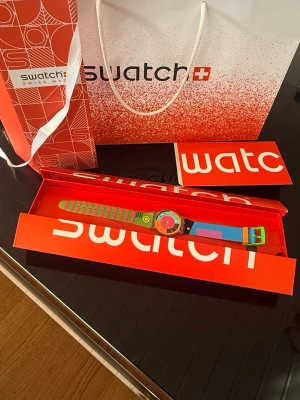 SWATCH Neon Hot Racer - Säljer en riktigt färgglad och unik Swatch klocka med randigt band i grönt och rosa samt detaljer i blått, orange och gult. Urtavlan är transparent med flera färgblock och cool design. Perfekt för dig som vill sticka ut med din stil!