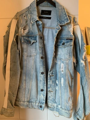 Ljusblå distressed jeansjacka Zara - Trendig ljusblå jeansjacka från Zara med slitna detaljer och råa kanter. Jackan har vita ränder längs ärmarna och två bröstfickor med lock. Oversized-normal passform och cool streetstyle-vibe. Perfekt för dig som vill sticka ut med en unik look.
