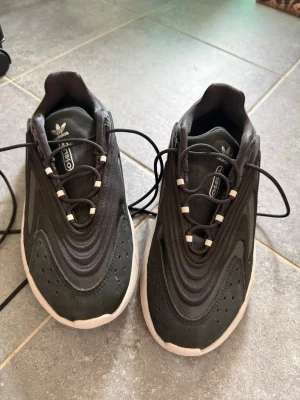 Svarta Adidas sneakers med vit sula - Säljer ett par svarta Adidas sneakers med vit sula och coola vågformade detaljer på ovandelen. Skorna har svarta snören med vita detaljer och Adidas-logga på plösen. Materialet är en mix av syntet och textil för en modern look.