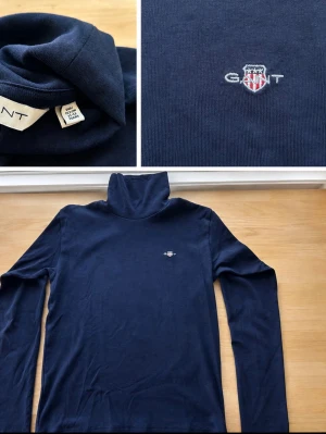 Mörkblå polotröja från Gant - Säljer en mörkblå polotröja från Gant med broderad logga på bröstet. Tröjan har lång ärm och hög polokrage, perfekt för lager på lager. Aldrig använt som ny