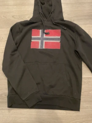 Svart hoodie med norsk flagga - Säljer en svart hoodie med stor norsk flagga tryckt på bröstet. Hoodien har huva med dragsko och en klassisk magficka framtill. Tillverkad i mjukt bomullsmaterial och har ribbade muddar vid ärmslut och nederkant. Perfekt för dig som gillar enkel och stilren design.