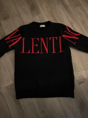 Svart stickad tröja med röd VALENTINO-text - Snygg svart stickad tröja från Valentino med stor röd text över bröstet och axlarna. Tröjan har rund halsringning och långa ärmar. Materialet ser ut att vara mjukt och passar perfekt för dig som vill ha en statement-piece i garderoben.