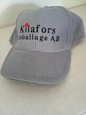 Grå keps med Kilafors-tryck - Snygg grå keps med broderad text 'Kilafors Emballage AB' framtill och en röd detalj ovanför K:et. Klassisk böjd skärm och justerbar baksida. Tillverkad i slitstarkt tyg, perfekt för en casual look.