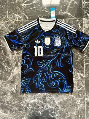 Argentina Adidas fotbollströja svart/blå - Snygg svart fotbollströja från Adidas med blått och vitt mönster, nummer 10 på bröstet och officiella Argentina-emblemet samt FIFA 2022-märke. Tröjan har vita ränder på axlarna och rund halsringning. Perfekt för dig som älskar fotboll och vill sticka ut på planen.