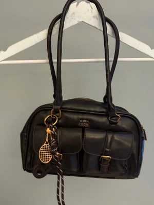 Brun handväska med fickor och detaljer - Snygg svart handväska  i läderlook med två framfickor med spännen, guldfärgade detaljer och dubbla axelband. Väskan har dragkedja upptill och en rolig nyckelring med tennisracket och boll som dekoration.
