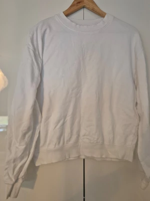 Vit basic sweatshirt - En enkel vit sweatshirt med rund hals och långa ärmar. Tröjan har ribbade muddar vid ärmslut och nederkant. Perfekt för en clean och avslappnad stil. Materialet känns mjukt och bekvämt, troligen bomull eller bomullsblandning.