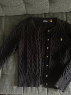 Svart kabelstickad kofta Polo Ralph Lauren - Kabelstickad svart kofta från Polo Ralph Lauren med rund halsringning och knappar framtill. Klassisk broderad logga på bröstet och ribbade muddar vid ärmslut och nederkant. Perfekt för lager på lager och snygg till jeans.