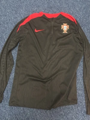 Portugal långärmad träningströja Nike - Snygg svart långärmad träningströja från Nike med Portugals landslagsmärke på bröstet och röda detaljer på axlarna. Tröjan har halv dragkedja framtill och är perfekt för fotbollsträning eller uppvärmning.